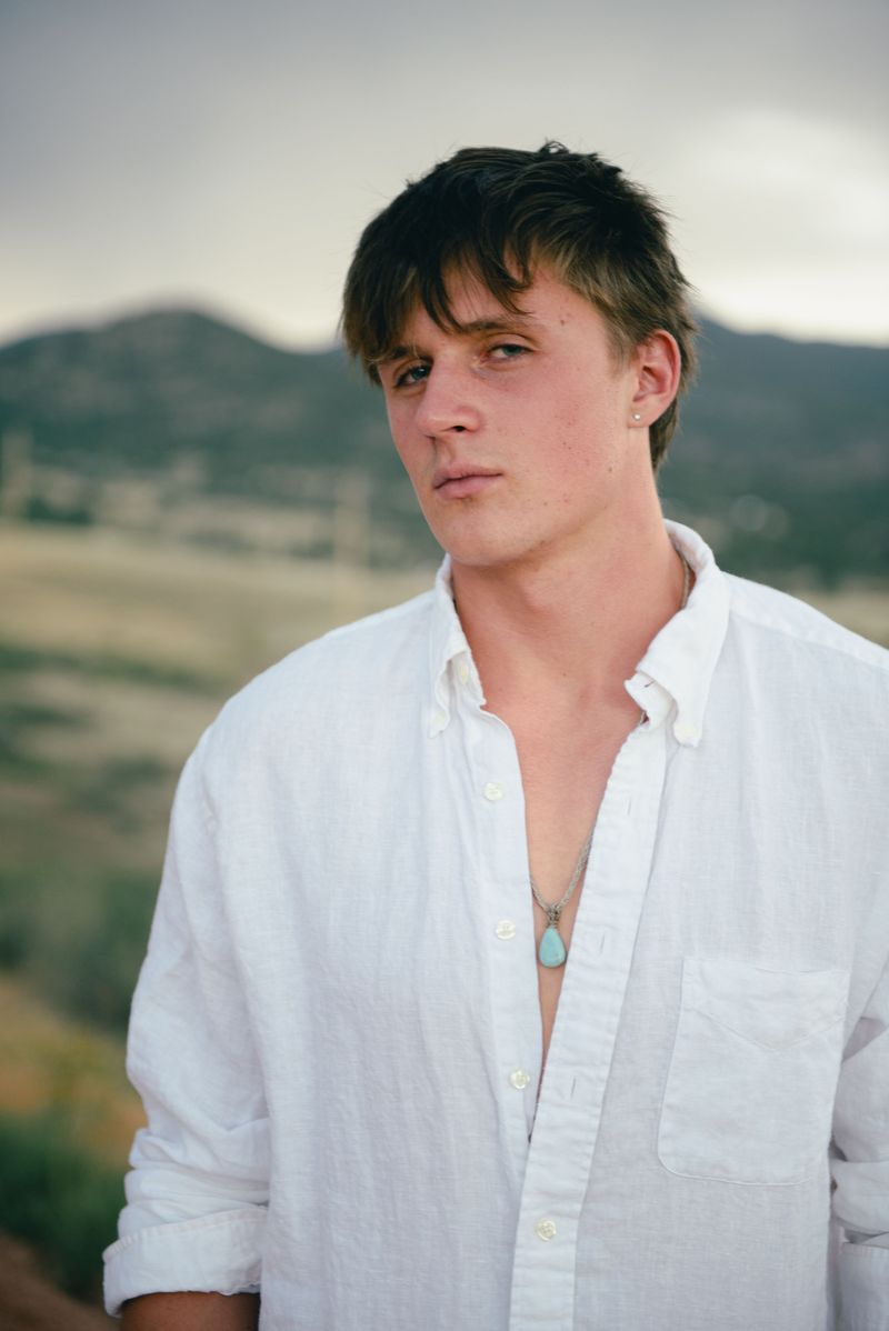 Portrait of a young man in an open white shirt, mountains softly blurred behind him.