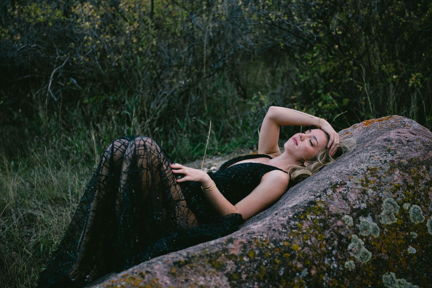 A young woman in a black dress reclines on a lichen-covered boulder in a dark forest.