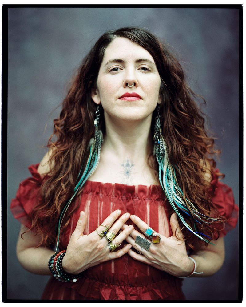 Portrait of Carolee Beckham in a red dress, hands at her heart, turquoise feathers in her hair.