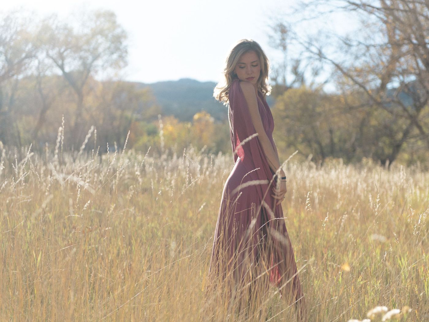 A young woman in a long red dress stands far off in a field of tall pale grass.