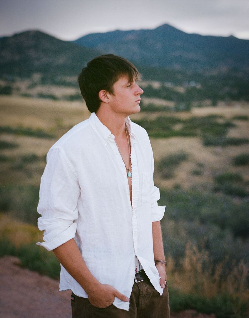 A young man in a white shirt stands before a mountain ridge at golden hour.