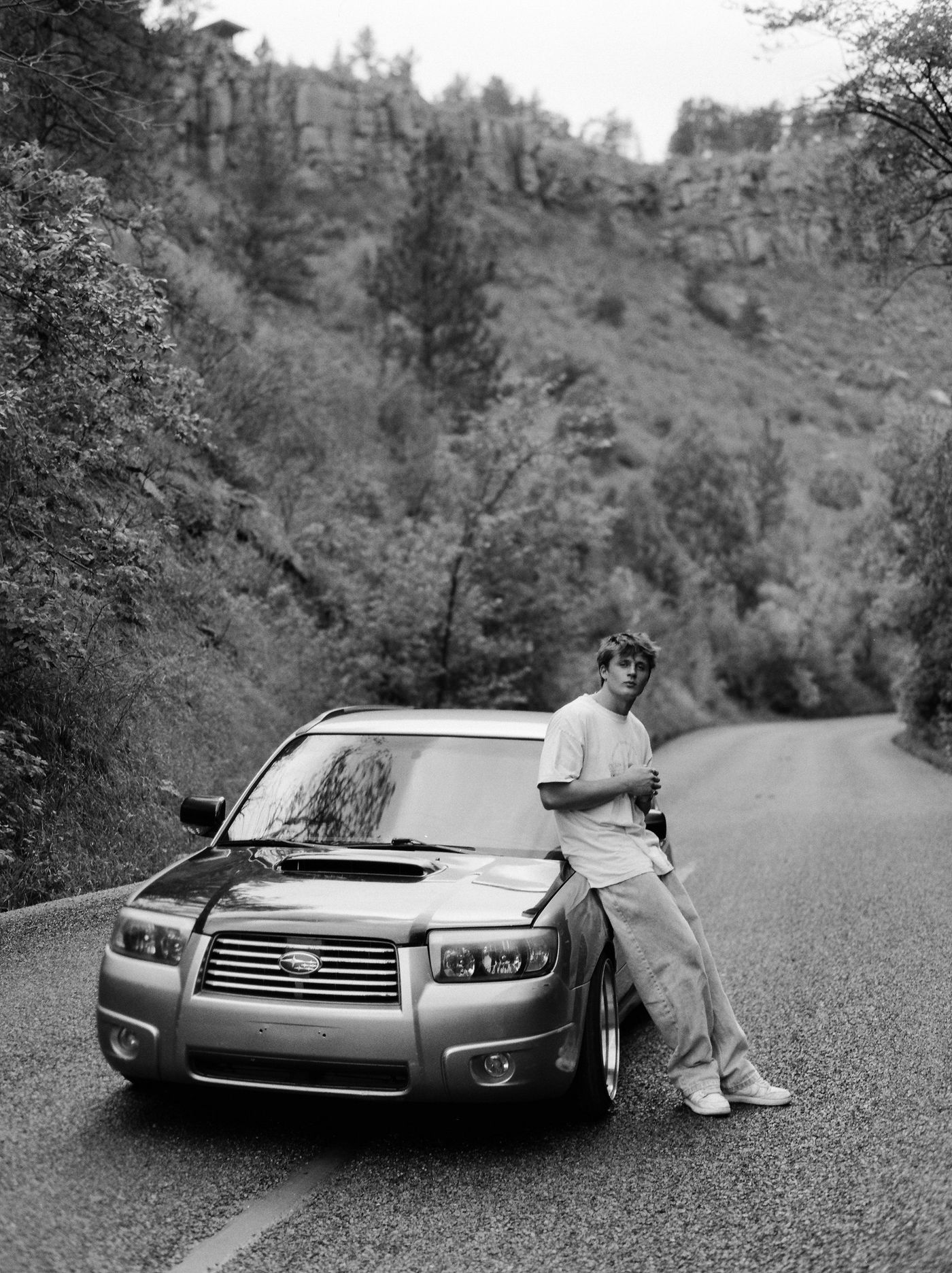 Black-and-white photo of a young man leaning on an SUV on a canyon road.