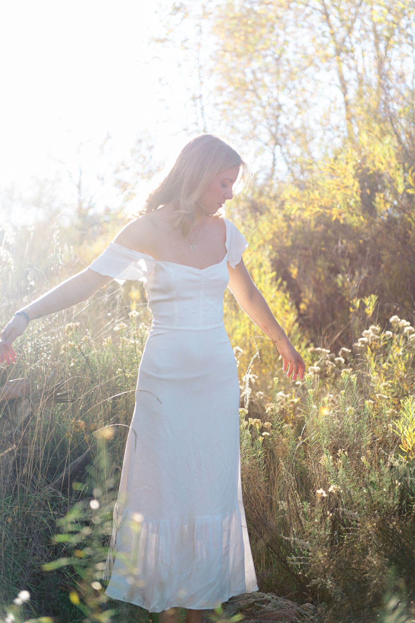 A young woman in a white dress opens her arms in a sunlit meadow.
