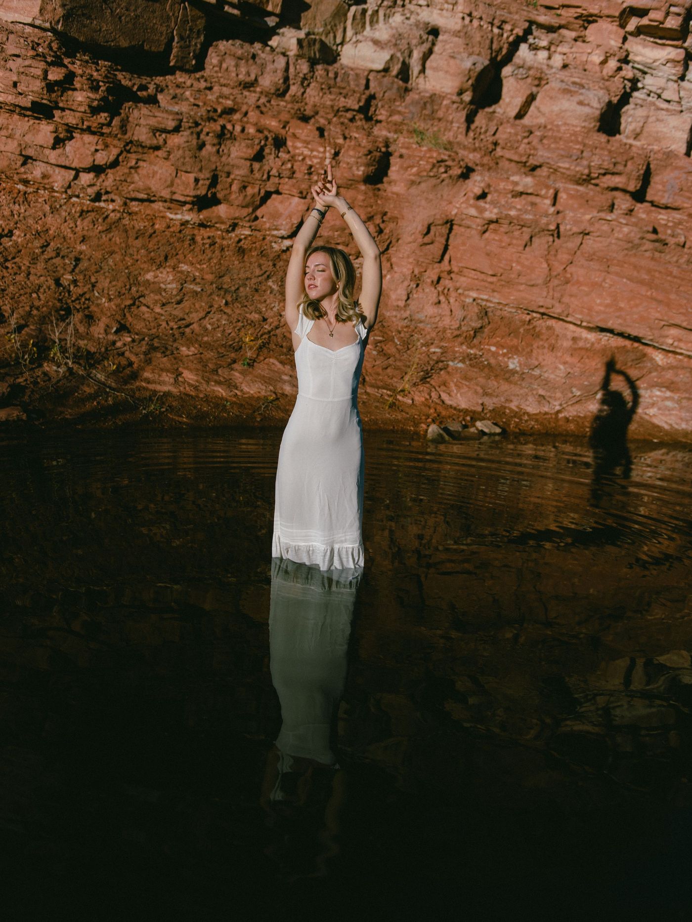 A young woman in white raises her arms beside red canyon rock and dark water.