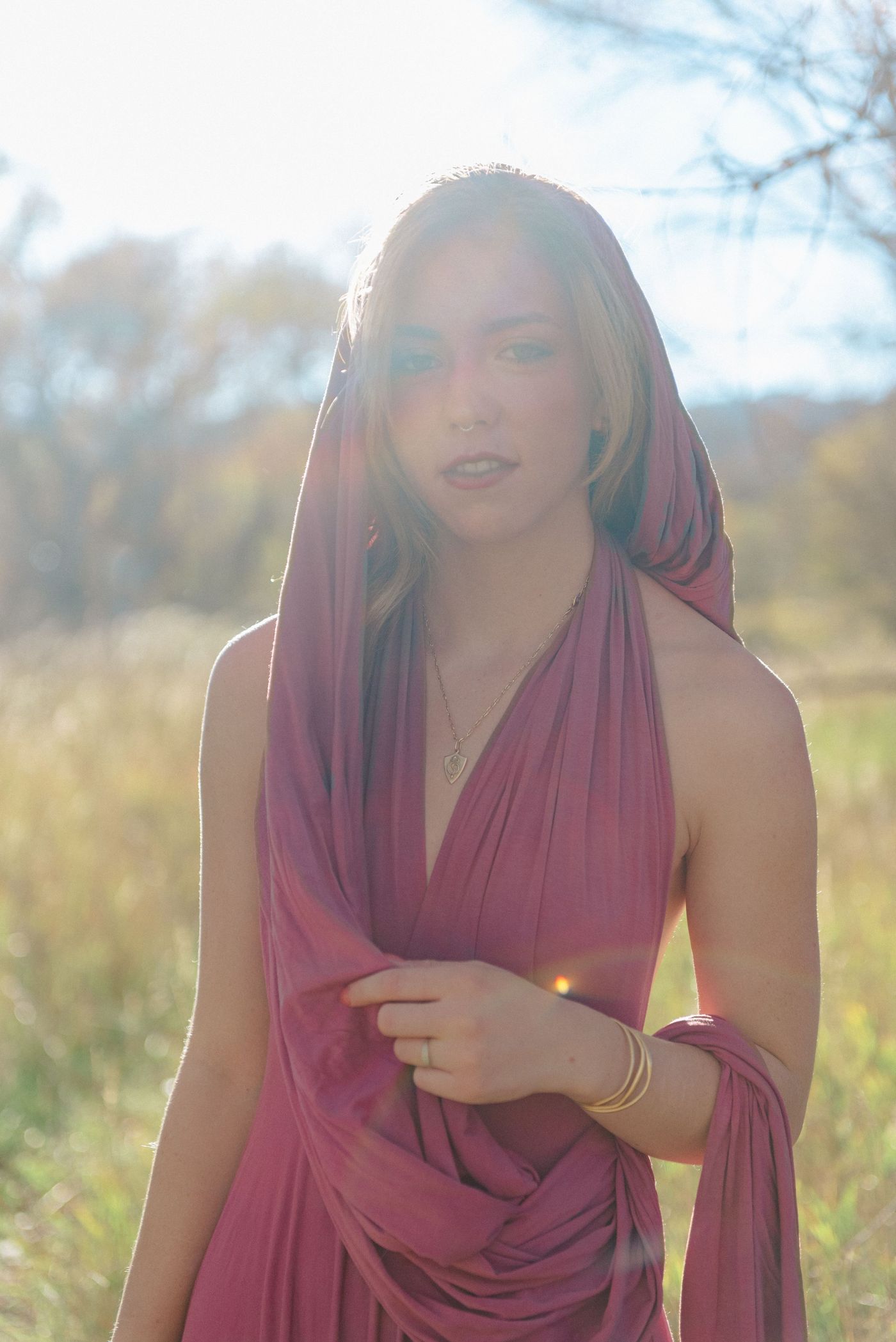 Close portrait of a young woman wrapped in a mauve hood and dress, backlit by the sun.