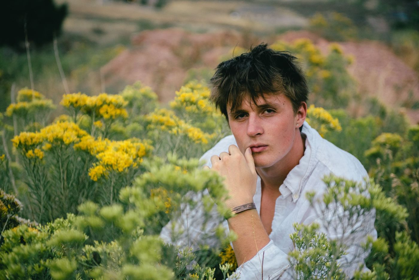 A young man in a white shirt sits among yellow wildflowers, chin resting on his hand.