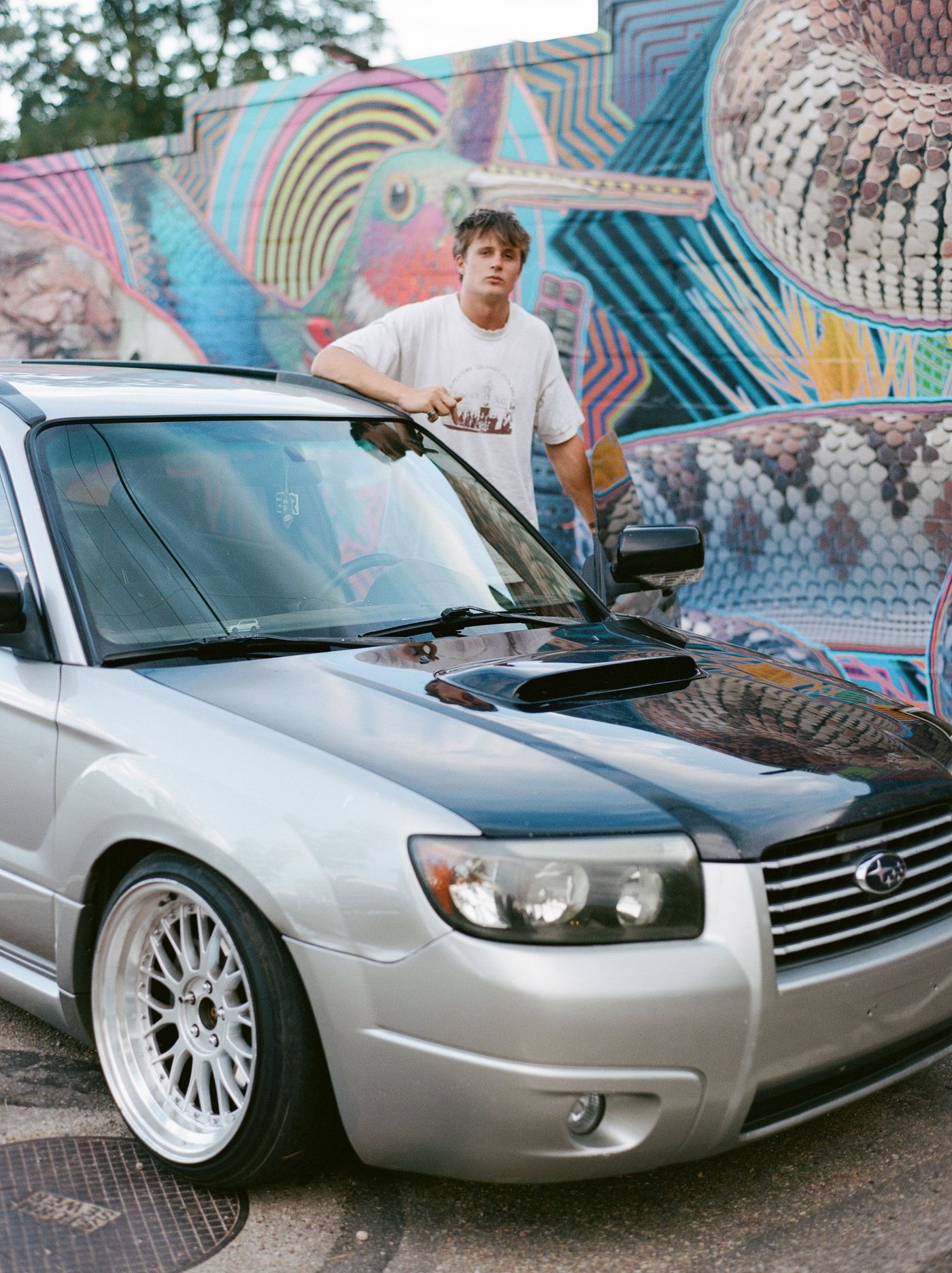 A young man sits on the hood of a silver SUV in front of a colorful mural.