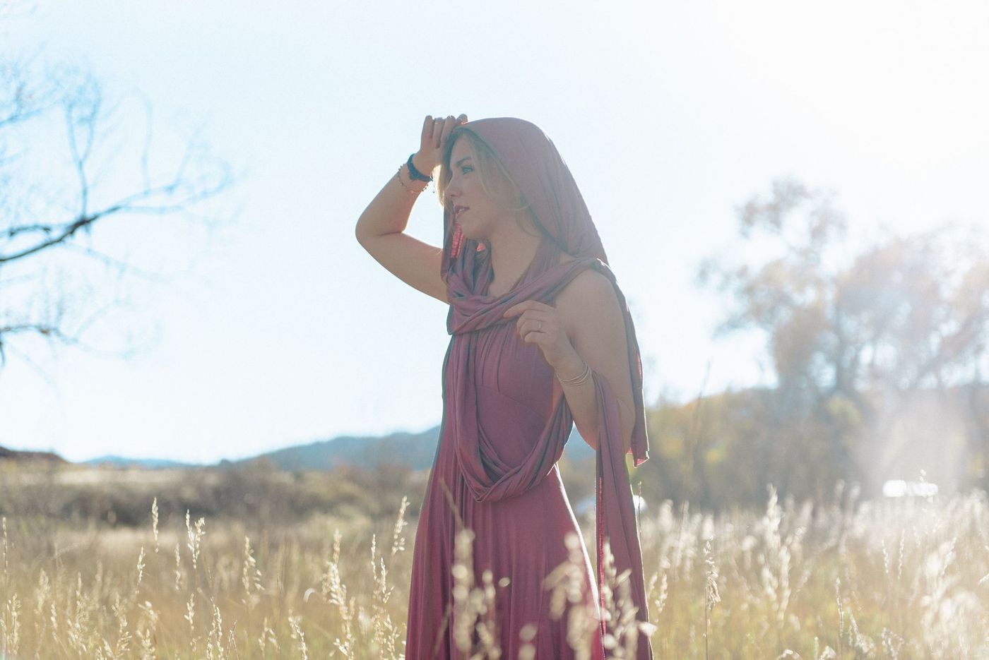 A young woman in a mauve dress stands in a meadow, hands lifted to her head.
