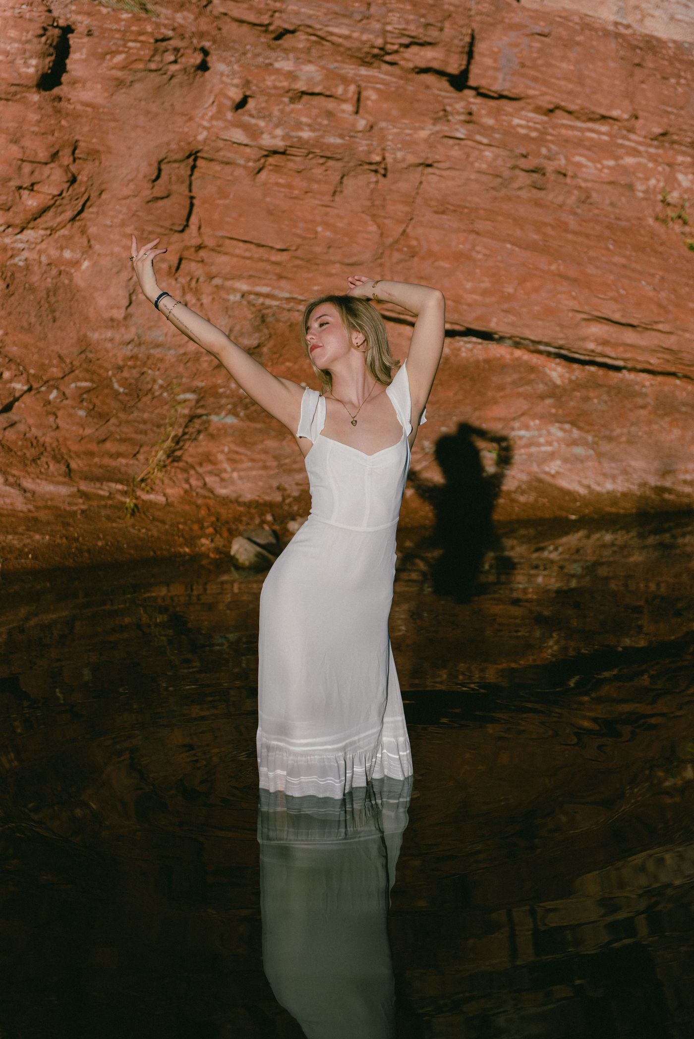 A young woman in white stands in a river beneath red rock cliffs, arms lifted overhead.