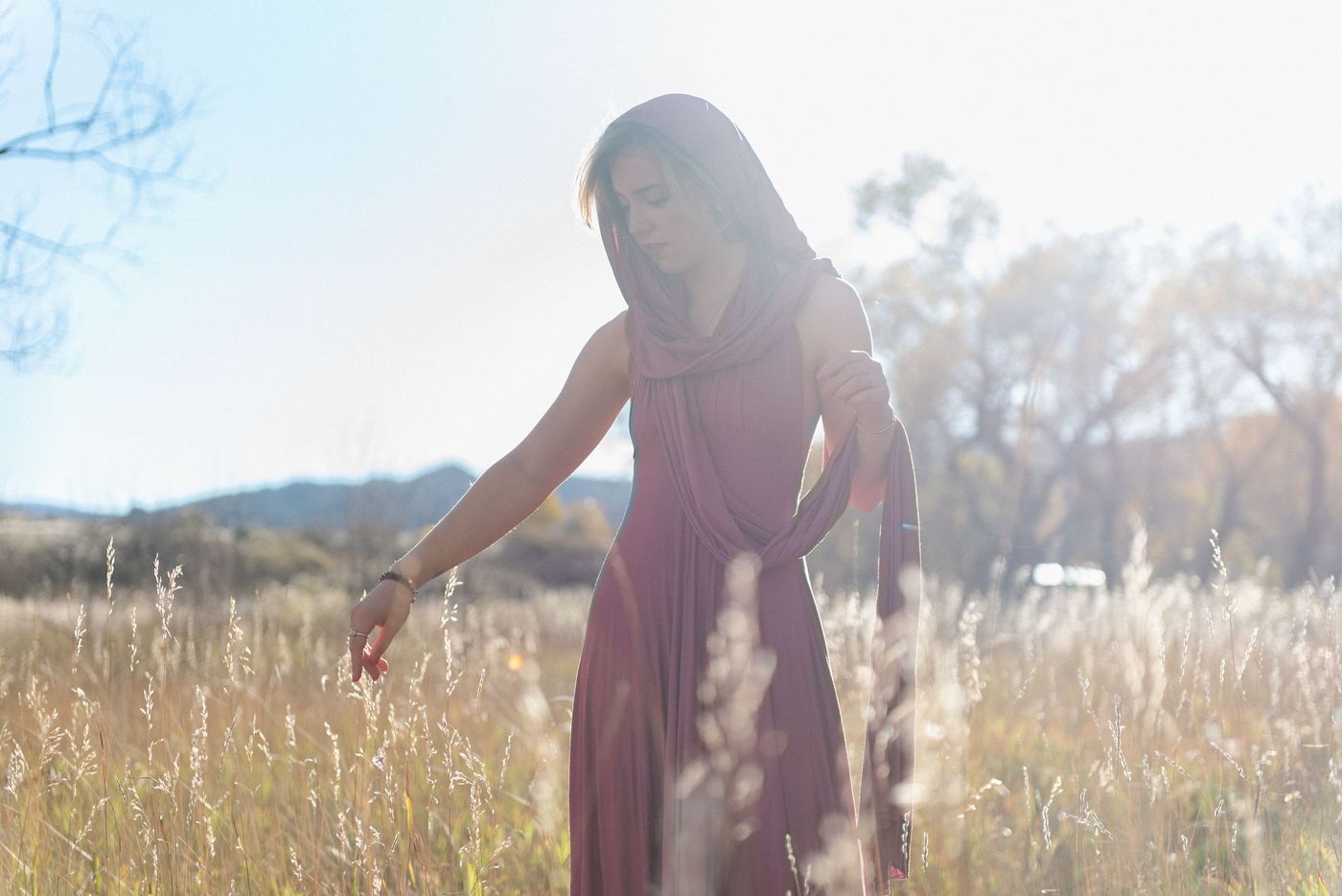 A young woman in a mauve dress walks through tall golden grass, one hand trailing through the stalks.