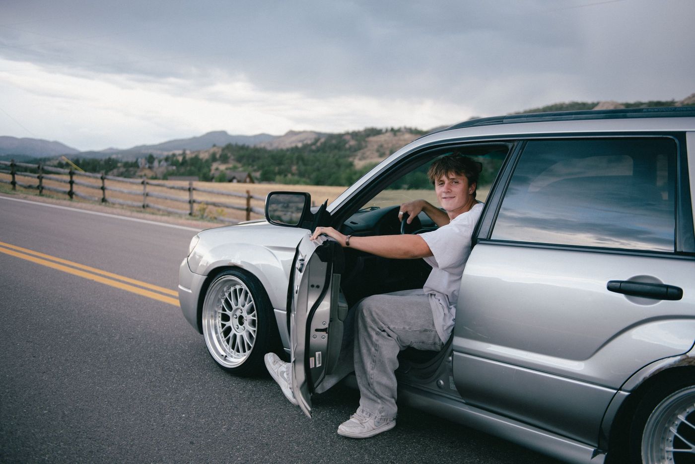A young man sits in the open door of a silver SUV parked on a country road.
