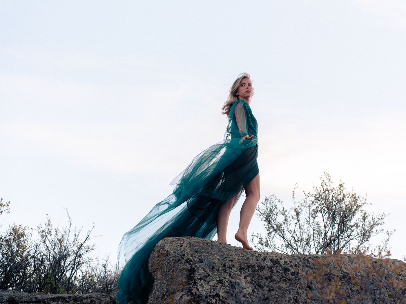 A young woman in a flowing teal gown stands on a rock as the fabric lifts in the wind.