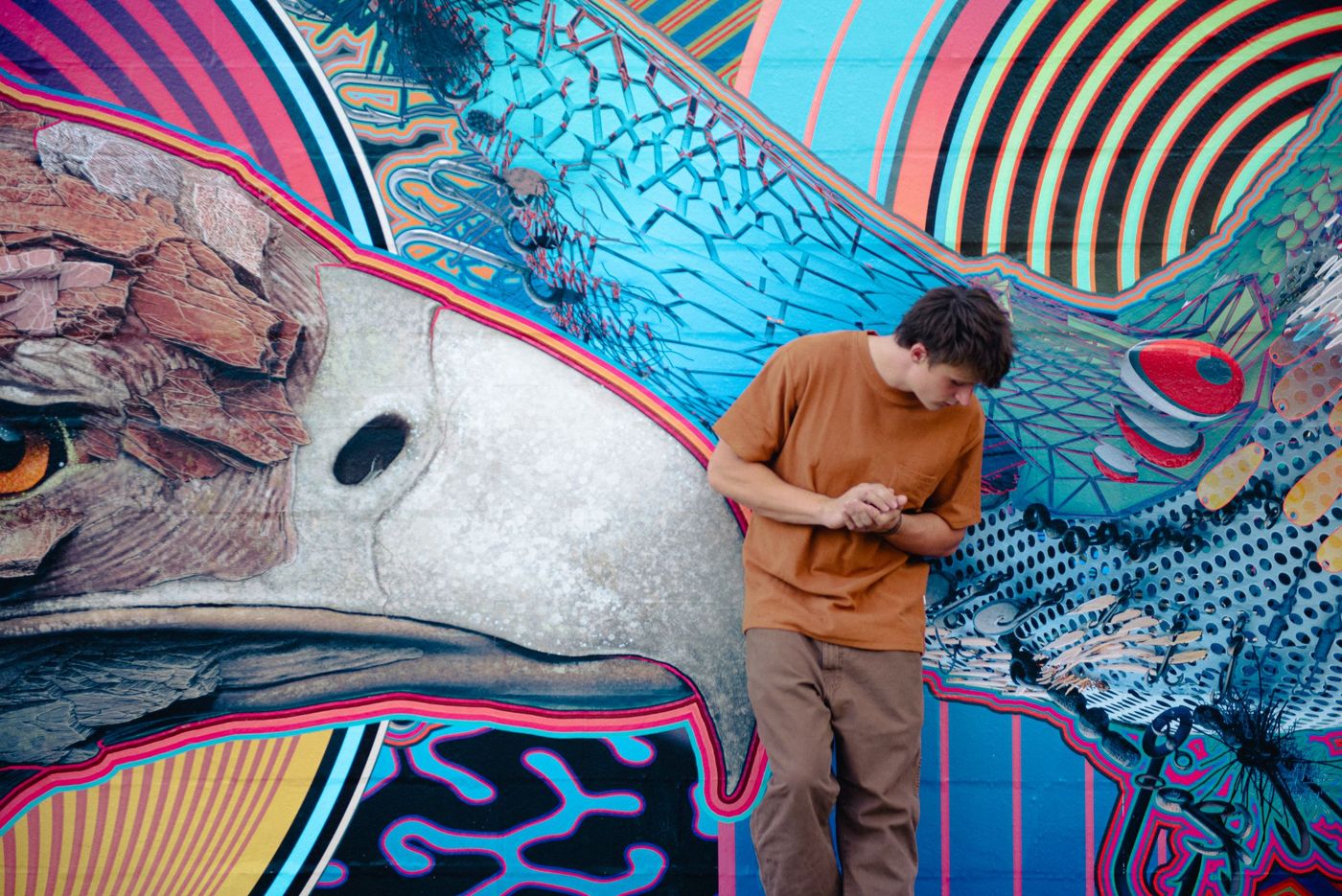 A young man in an orange shirt leans against a vivid mural of an eagle.